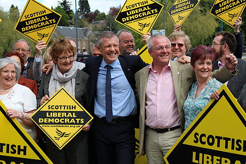 Alt text: Willie Rennie MSP with Cllr Peter Barrett and Lib Dem campaigners in Perth. The crowd are holing large gold Lib Dem diamond signs. Willie and Peter are smiling and looking to their left.  Willie  and 